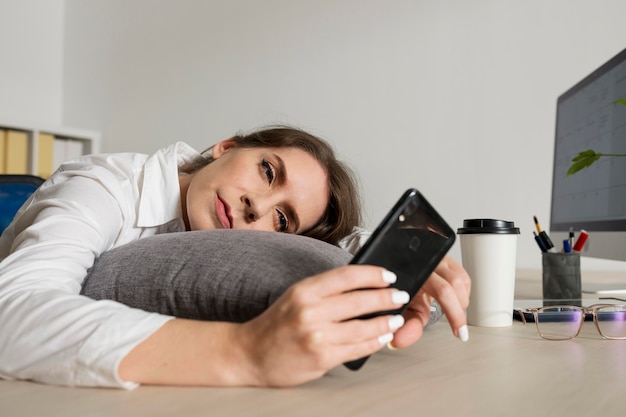 Profissional com a cabeça apoiada na mesa olhando o celular, comportamento associado à autossabotagem no trabalho